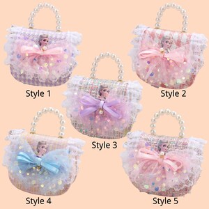 K&ouml;nnte beinhalten: F&uuml;nf kleine, dekorative Handtaschen mit Perlenhenkeln. Jede Handtasche hat eine Charakterfigur, eine Schleife und Spitzenbesatz. Die Handtaschen sind in verschiedenen Farben erh&auml;ltlich, darunter Rosa, Blau und Lila, mit dem Text "Style" und einer Nummer.
