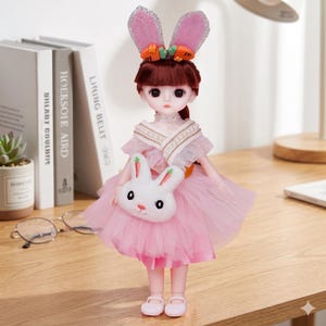 May include: A doll with brown hair wearing a pink tulle dress, a white rabbit-shaped crossbody bag, and a headband featuring pink glittery rabbit ears with small orange carrot accents. The doll has large dark eyes and wears light pink shoes.
