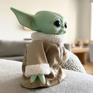 May include: A green-skinned doll with large black eyes and pointed ears, dressed in a tan coat with a white, fluffy collar and cuffs. The doll is a character from a popular science fiction franchise.