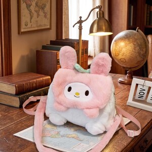 May include: A soft white and pink My Melody crossbody bag featuring a cartoon face and bunny ears. The bag has a long pink strap and is placed on a map. The bag is on a wooden desk with a lamp and globe in the background.