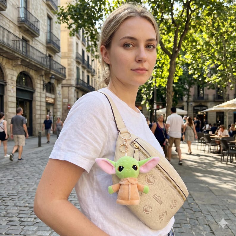 May include: A plush green and tan character keychain is attached to a cream-colored crossbody bag. The bag has a gold zipper and a gold chain. The person is wearing a white t-shirt.