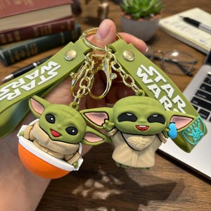 Puede incluir: Un juego de dos llaveros con figuras de silicona verde del personaje Grogu de Star Wars. Una figura est&aacute; sentada en un cuenco naranja, mientras que la otra est&aacute; de pie con un accesorio azul. Ambas est&aacute;n unidas a correas verde oliva con texto Star Wars.