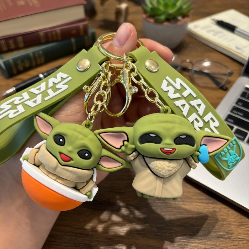 Puede incluir: Un juego de dos llaveros con figuras de silicona verde del personaje Grogu de Star Wars. Una figura est&aacute; sentada en un cuenco naranja, mientras que la otra est&aacute; de pie con un accesorio azul. Ambas est&aacute;n unidas a correas verde oliva con texto Star Wars.