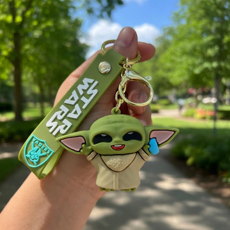 Puede incluir: Un llavero de silicona verde con un dije del personaje Grogu con orejas grandes y t&uacute;nica beige, unido a una correa verde a juego con el texto blanco STAR WARS y un peque&ntilde;o icono azul de Vader. Incluye una anilla de metal de tono dorado.