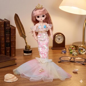 May include: A doll with pink hair and a gold crown, wearing a white off-the-shoulder top with a blue shell pendant and a pink sequined mermaid tail skirt featuring a layered, multi-colored tulle train that flares out at the base.