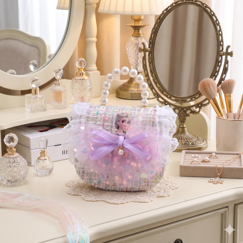 K&ouml;nnte beinhalten: Eine kleine, runde Handtasche mit Perlenhenkel und dekorativer Schleife. Die Tasche ist mit Pailletten und einer Puppenfigur verziert. Die Tasche steht auf einer wei&szlig;en Oberfl&auml;che mit anderem Sch&ouml;nheitszubeh&ouml;r.