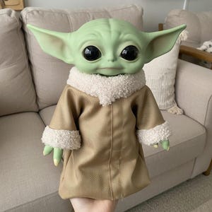 May include: A green-skinned doll with large black eyes and pointed ears, dressed in a tan coat with white fluffy trim. The doll's hands and feet are green with yellow nails. The doll is a character from a popular science fiction franchise.