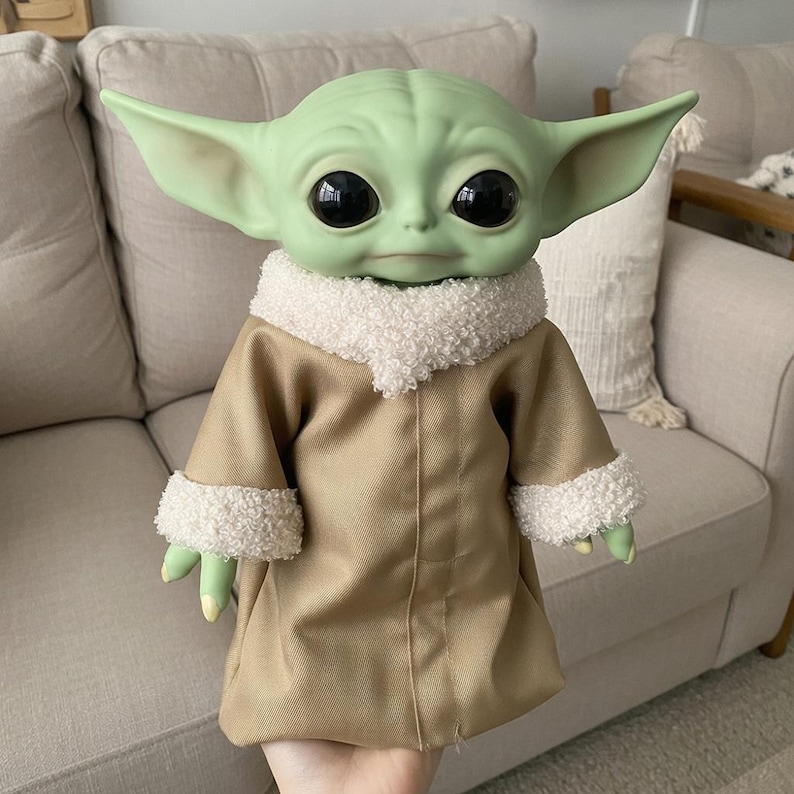 May include: A green-skinned doll with large black eyes and pointed ears, dressed in a tan coat with white fluffy trim. The doll's hands and feet are green with yellow nails. The doll is a character from a popular science fiction franchise.