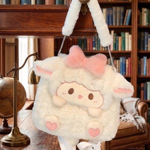 May include: A cream-coloured sheep-shaped handbag with a pink bow and heart details. The bag has a cartoon sheep face and a white strap. The bag is suspended from a wooden door.