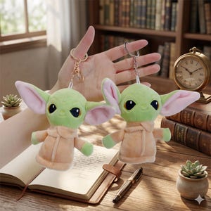 May include: Two plush green character keychains with large ears and tan robes. The keychains are attached to gold-colored metal rings. The background includes a desk with a book, pen, and small potted plant.