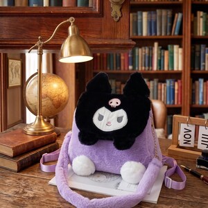 May include: A lavender plush backpack featuring a black character face and white details. The bag has a long, fluffy strap and two white pom-poms. The bag is on a wooden surface.
