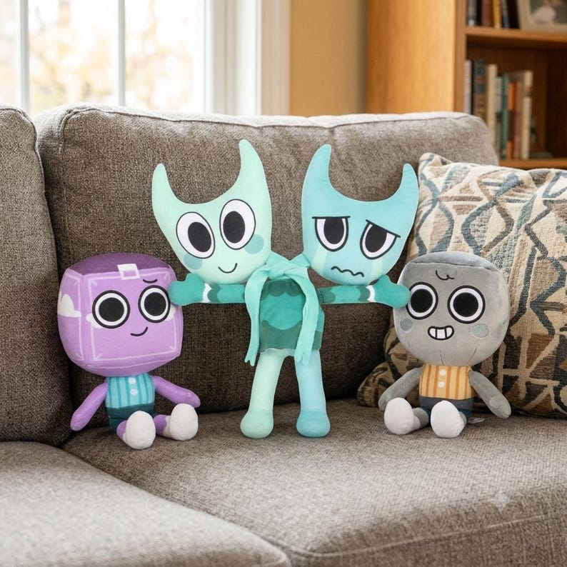 May include: Three plush toys featuring cartoon characters. On the left, a purple cube-shaped character with white accents. In the centre, a two-headed light blue character with a tied ribbon. On the right, a round grey character with a striped shirt.