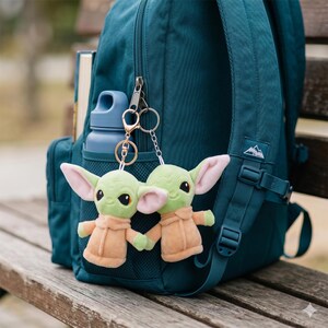 May include: Two plush green alien keychains with large ears and tan robes hang from a teal backpack. A blue water bottle and a book are in the backpack's pockets. The backpack is on a wooden bench.