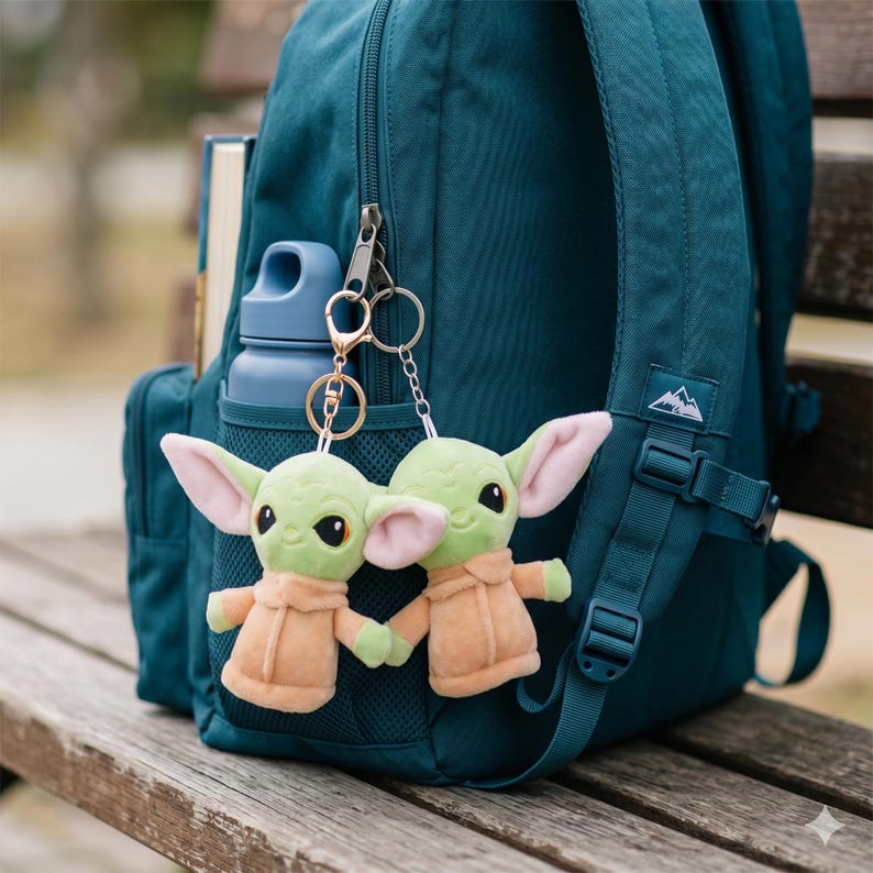May include: Two plush green alien keychains with large ears and tan robes hang from a teal backpack. A blue water bottle and a book are in the backpack's pockets. The backpack is on a wooden bench.
