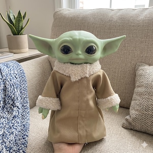 May include: A green-skinned doll with large black eyes and oversized ears, wearing a tan coat with a fluffy white collar and cuffs. The doll is a character from a popular science fiction franchise.