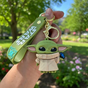 Puede incluir: Un llavero de silicona con un personaje verde de orejas grandes, vestido con una t&uacute;nica beige y sosteniendo una peque&ntilde;a mariposa azul. Incluye una correa verde con el texto blanco STAR WARS y un escudo con la palabra VADERS.