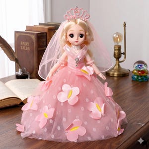 May include: A doll with blonde hair wearing a pink tiara and a pink veil. The doll is dressed in a pink gown featuring a polka dot tulle overlay and several pink flower appliqu&eacute;s with yellow centers. The doll has large eyes and a white collar.