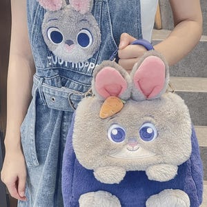 May include: A fluffy bunny-shaped handbag in blue and grey, with pink inner ears, large blue eyes, and a small orange carrot. A matching bunny face is on the denim dungarees.