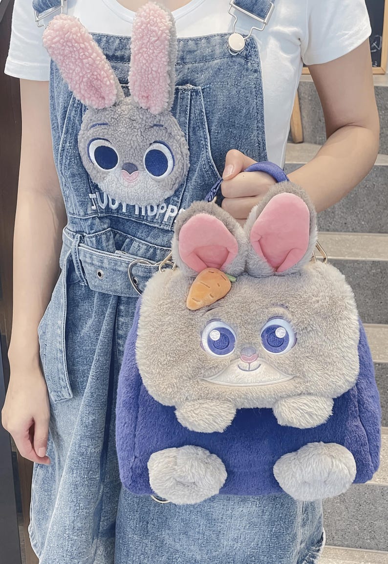 May include: A fluffy bunny-shaped handbag in blue and grey, with pink inner ears, large blue eyes, and a small orange carrot. A matching bunny face is on the denim dungarees.
