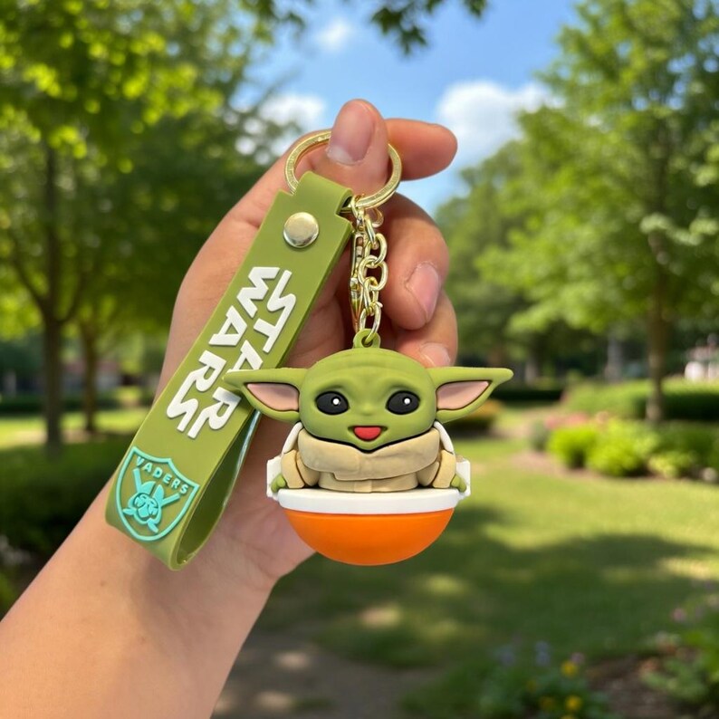 Puede incluir: Un llavero de silicona verde con una figura del personaje Grogu sentado en una c&aacute;psula naranja y blanca. La correa verde adjunta muestra el texto blanco STAR WARS y un logotipo azul claro con la palabra VADERS sobre un gr&aacute;fico de casco.
