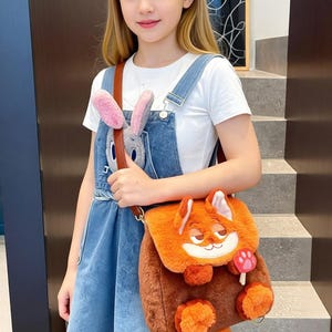 May include: A fluffy orange and brown fox-shaped backpack with a mushroom detail. The backpack has a brown strap and is worn over the shoulder. A grey bunny plush is attached to the denim dungarees.