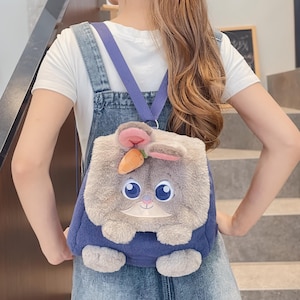 May include: A novelty backpack in the shape of a cartoon rabbit. The backpack has a fluffy grey and white bunny face with large blue eyes, pink ears, and a small orange carrot. It has blue straps and a blue body.