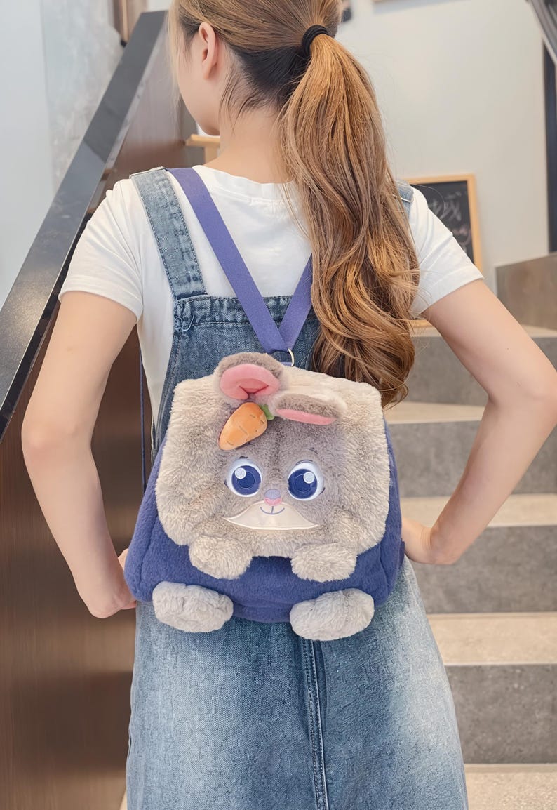 May include: A novelty backpack in the shape of a cartoon rabbit. The backpack has a fluffy grey and white bunny face with large blue eyes, pink ears, and a small orange carrot. It has blue straps and a blue body.