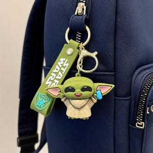 Puede incluir: Una mochila azul marino con un llavero de Star Wars que incluye una figura verde de Grogu y una correa de silicona verde a juego con el texto STAR WARS impreso en blanco, sujeto al tirador de la cremallera con una anilla met&aacute;lica dorada.