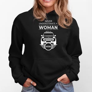 May include: Black hooded sweatshirt featuring the text "NEVER UNDERESTIMATE A WOMAN WITH A MINI" in white, along with a white Mini Cooper car graphic. The sweatshirt has a front pocket and a drawstring hood.