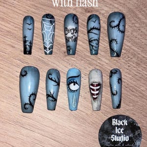 May include: A set of ten press-on nails with a light blue base and black hand-painted designs including spiderwebs, thorny vines, a crying eye, and the text 'Corps Bride'. The nails feature a glossy finish and are arranged in two rows.