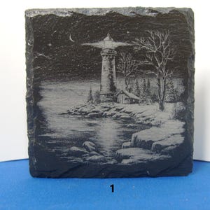 May include: A square black slate coaster featuring a laser-engraved white illustration of a lighthouse on a rocky shore at night. The scene includes a crescent moon, a small cabin, bare trees, and a reflection of the lighthouse beam on the water.