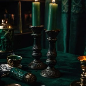May include: Two dark green pillar candles with lit flames stand in ornate, dark metal candle holders. A deck of illustrated cards, a small green bowl, a cylindrical object with gold accents, and a metal lid are arranged on a dark green surface.