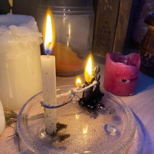 May include: A tall white taper candle and a small black candle are connected by a piece of twine on a clear glass plate. Both candles are lit, producing bright yellow flames. The white candle stands upright, while the black candle is shorter.