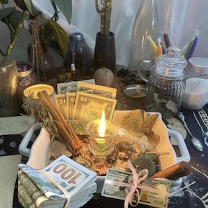 May include: A white square ceramic dish containing a lit green candle, cinnamon sticks, assorted coins, and paper currency. A thick stack of one hundred dollar bills is secured with a rubber band, and a single bill is tied with a light pink ribbon.