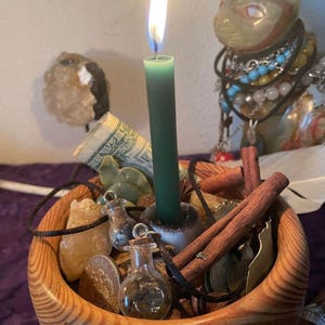 May include: A wooden bowl containing a lit dark green taper candle, cinnamon sticks, small glass vials with cork stoppers, a rolled one-dollar bill, a metallic coin, and yellow crystals. The bowl has a visible wood grain pattern and a rounded shape.