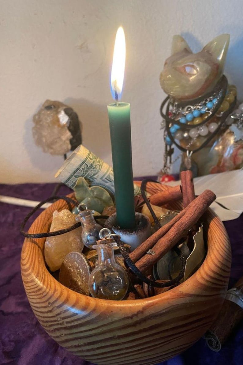 May include: A wooden bowl containing a lit dark green taper candle, cinnamon sticks, small glass vials with cork stoppers, a rolled one-dollar bill, a metallic coin, and yellow crystals. The bowl has a visible wood grain pattern and a rounded shape.