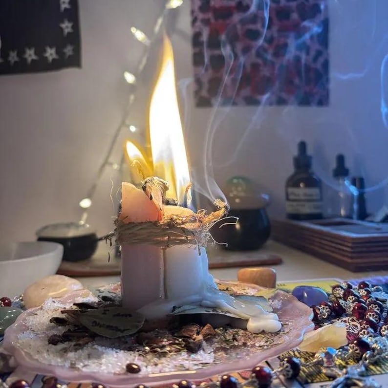 May include: A burning candle with a tall, bright flame and rising smoke, wrapped in twine. The candle sits on a translucent pink plate surrounded by scattered dried herbs, small stones, and a string of dark red beads on a flat surface.