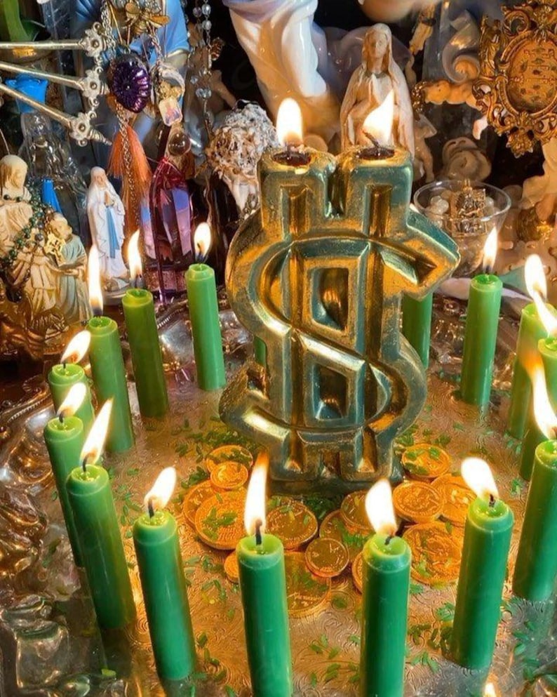 May include: A large, metallic gold dollar sign candle with two lit wicks, surrounded by numerous thin, lit green taper candles and scattered gold-colored coins on a reflective silver tray, with various religious figurines in the background.