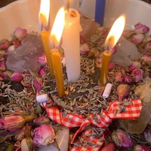 May include: A white bowl filled with dried pink rosebuds, lavender sprigs, and clear quartz crystals. Three lit candles, two yellow and one white, are positioned in the center. A red and white checkered ribbon is tied around the candle base.