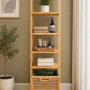 May include: A tall, light-brown bamboo shelf unit with multiple shelves. The shelves hold folded towels, toiletries, and a potted plant. A green fabric-lined basket is at the bottom. The unit is in a bathroom setting.