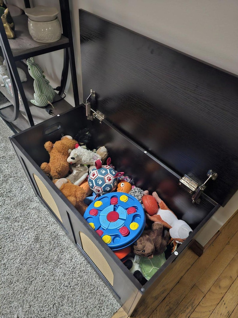 May include: A black wooden toy chest with a hinged lid, filled with various stuffed animals and toys. The chest has decorative side panels and is open, revealing the contents. The toys include teddy bears, balls, and a blue and red toy.