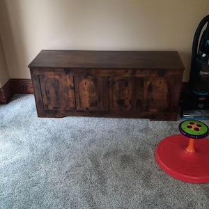 May include: A large, dark brown wooden chest with a rectangular shape. The chest has a flat top and arched panels on the front. It sits on a grey carpeted floor. A red and green toy and a vacuum cleaner are also visible.