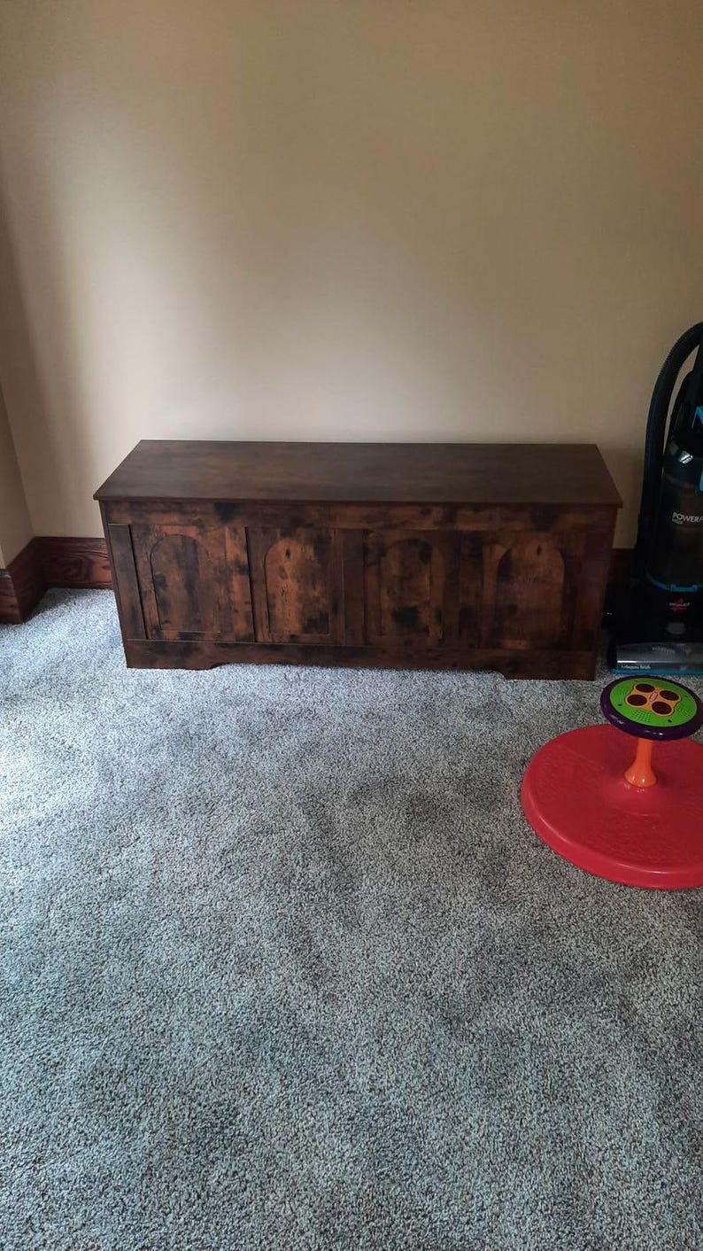 May include: A large, dark brown wooden chest with a rectangular shape. The chest has a flat top and arched panels on the front. It sits on a grey carpeted floor. A red and green toy and a vacuum cleaner are also visible.