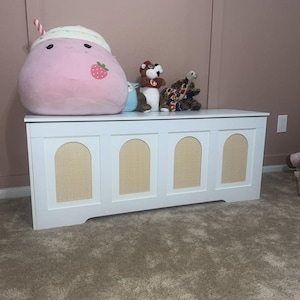 May include: A white wooden storage chest with four arched panels featuring woven cane inserts. The chest is topped with plush toys, including a large pink strawberry-themed plushie and several stuffed animals.