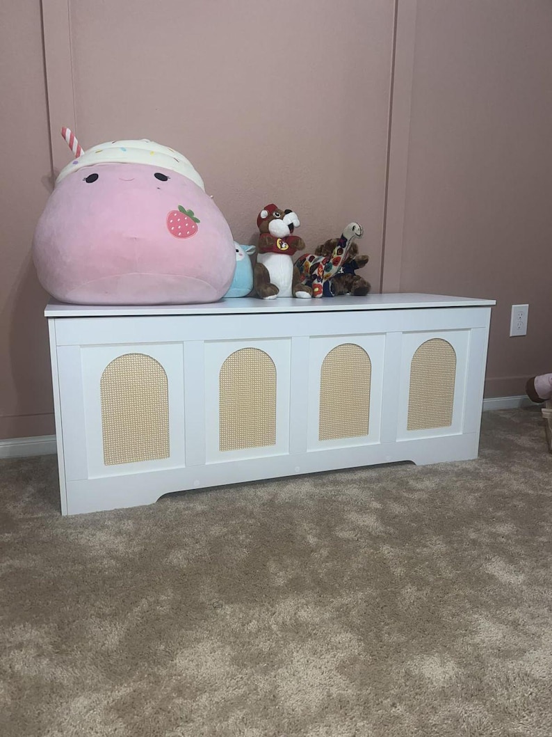 May include: A white wooden storage chest with four arched panels featuring woven cane inserts. The chest is topped with plush toys, including a large pink strawberry-themed plushie and several stuffed animals.