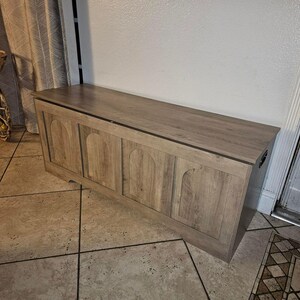 May include: A light brown wooden storage chest with a flat top and arched paneling on the front. The chest is rectangular and has a dark trim along the top edge. It is placed against a white wall.