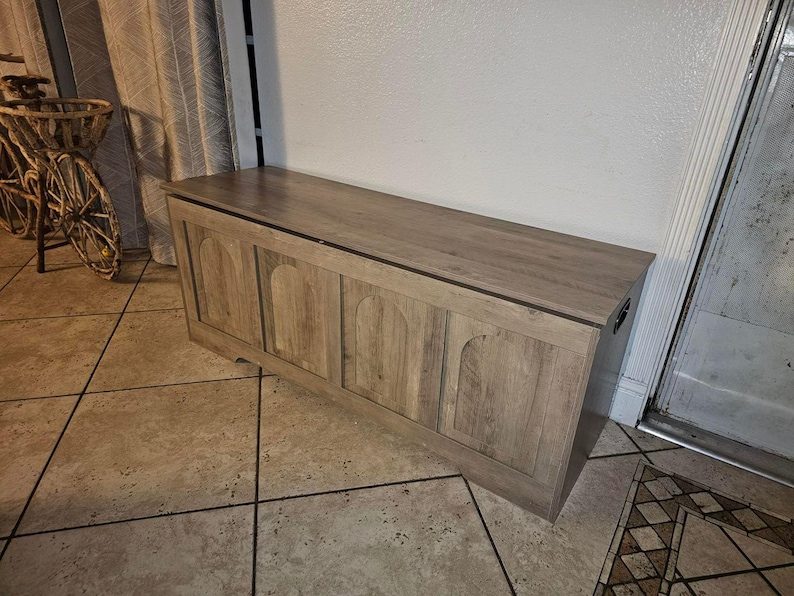 May include: A light brown wooden storage chest with a flat top and arched paneling on the front. The chest is rectangular and has a dark trim along the top edge. It is placed against a white wall.