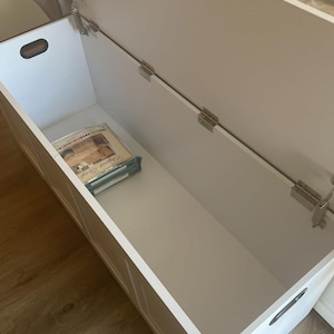 May include: A white wooden storage chest with the lid open, revealing an empty interior. The chest has a rectangular shape with a handle on the side. The lid is attached with metal hinges. A stack of items is visible inside the chest.