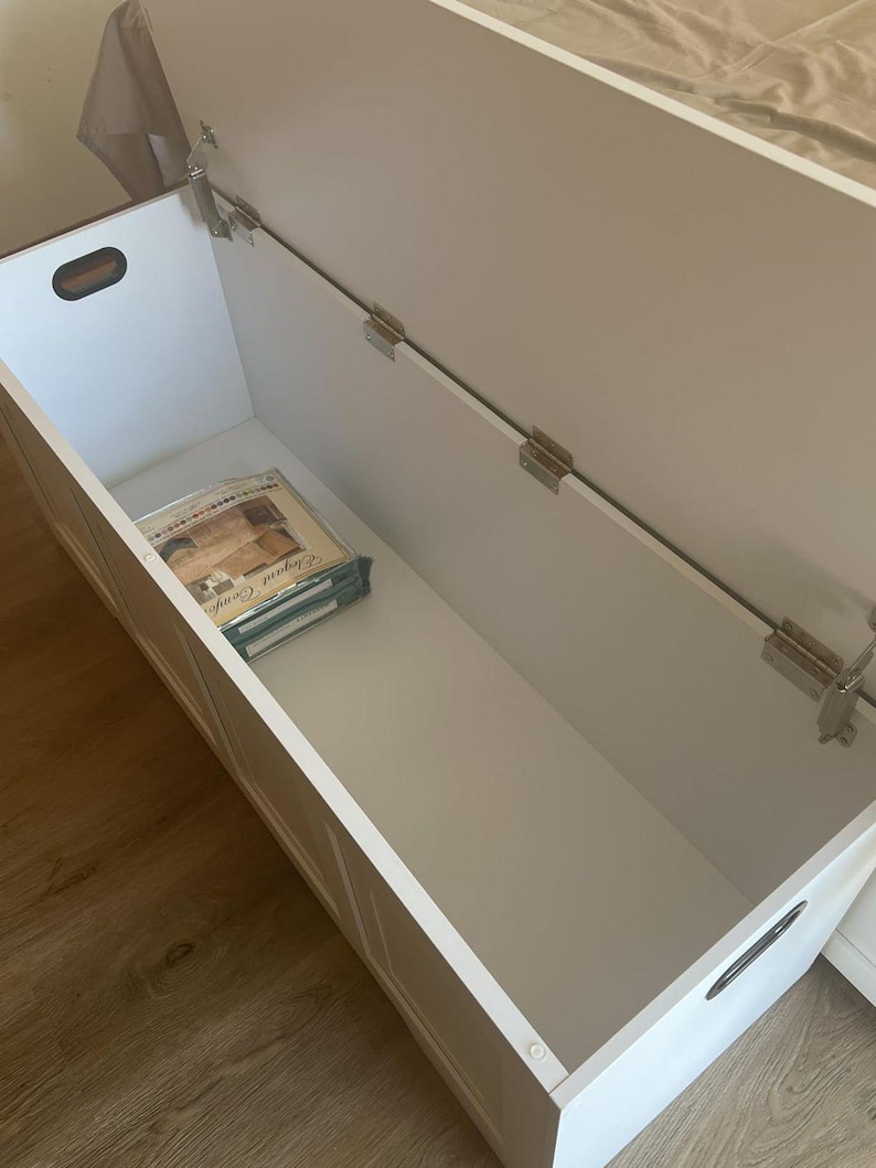 May include: A white wooden storage chest with the lid open, revealing an empty interior. The chest has a rectangular shape with a handle on the side. The lid is attached with metal hinges. A stack of items is visible inside the chest.