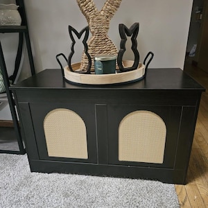 May include: A black storage cabinet with two arched rattan panels. A wooden tray on top holds a woven rabbit figure, two black bunny silhouettes, and a green candle. The cabinet sits on a grey carpet.